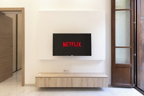 Un saló modern amb un televisor penjat que mostra 'NETFLIX' a la pantalla, una banca de fusta a sota i una porta a l'esquerra.