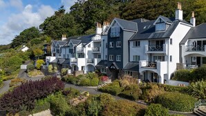 Une rangée de maisons modernes à plusieurs étages, dotées de balcons, entourées d’une végétation luxuriante et bordées d’une allée sinueuse.