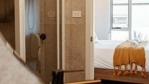 Ein Zimmer mit einer gläsernen Duschkabine, einem Bett mit gelber Decke und einem Fenster mit Tageslicht.