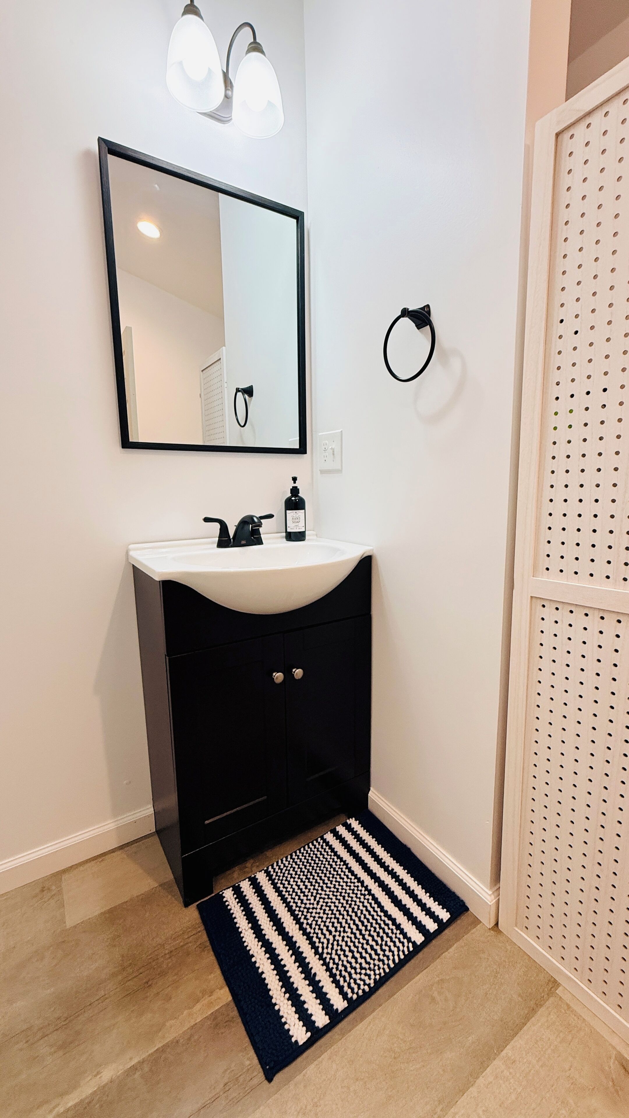 Une salle de bain avec un meuble-lavabo noir, un lavabo blanc et un miroir au-dessus. On y trouve un porte-serviettes noir fixé au mur et une porte blanche ornée d’un motif ajouré.
