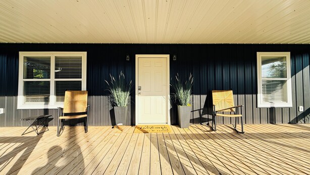A wooden deck with two wicker chairs, a door with a welcome mat, and potted plants.