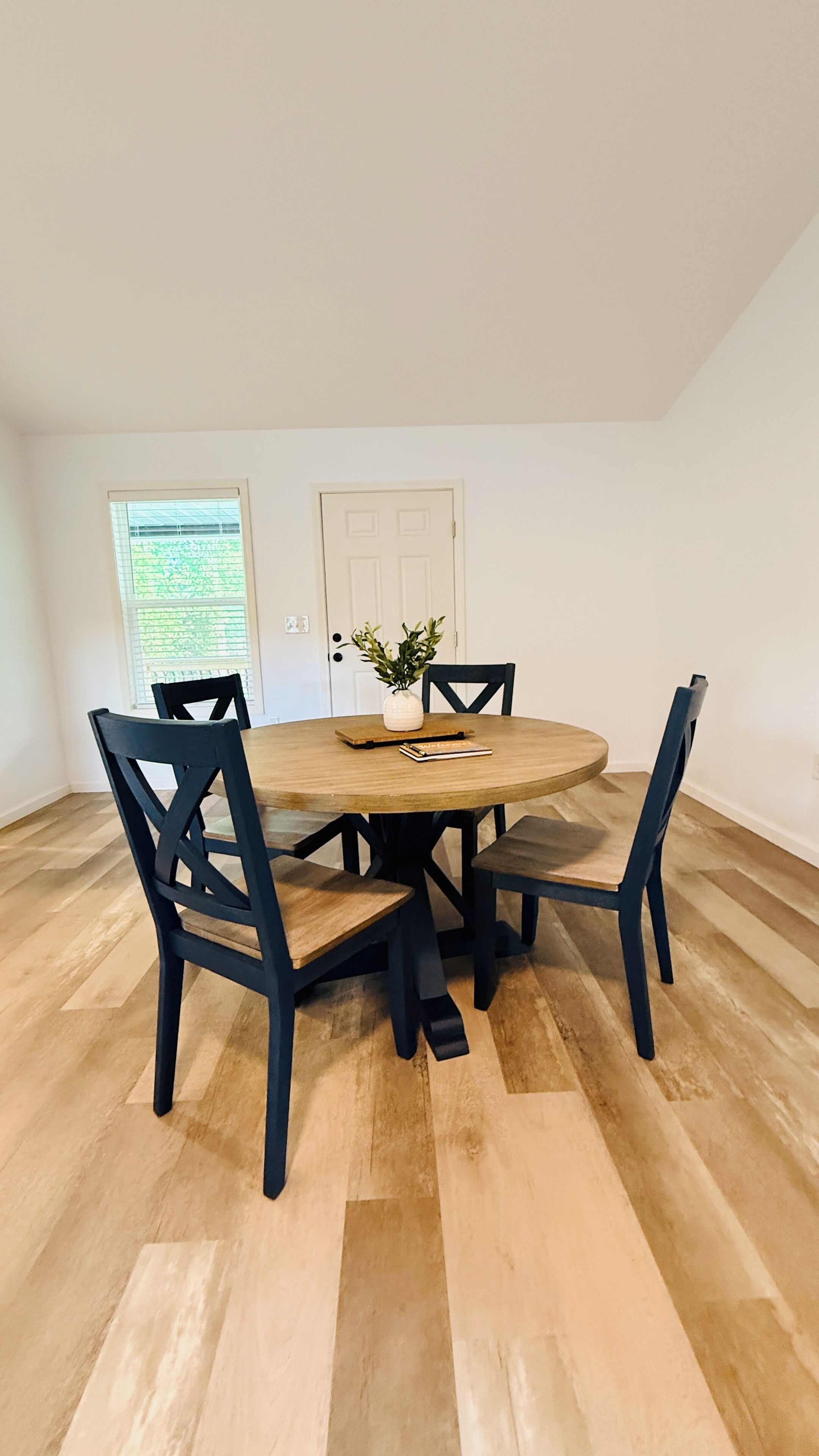 Un coin repas comprenant une table ronde en bois, quatre chaises noires, une porte blanche et une fenêtre avec des stores.