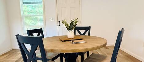 Un coin repas comprenant une table ronde en bois, quatre chaises noires, une porte blanche et une fenêtre avec des stores.