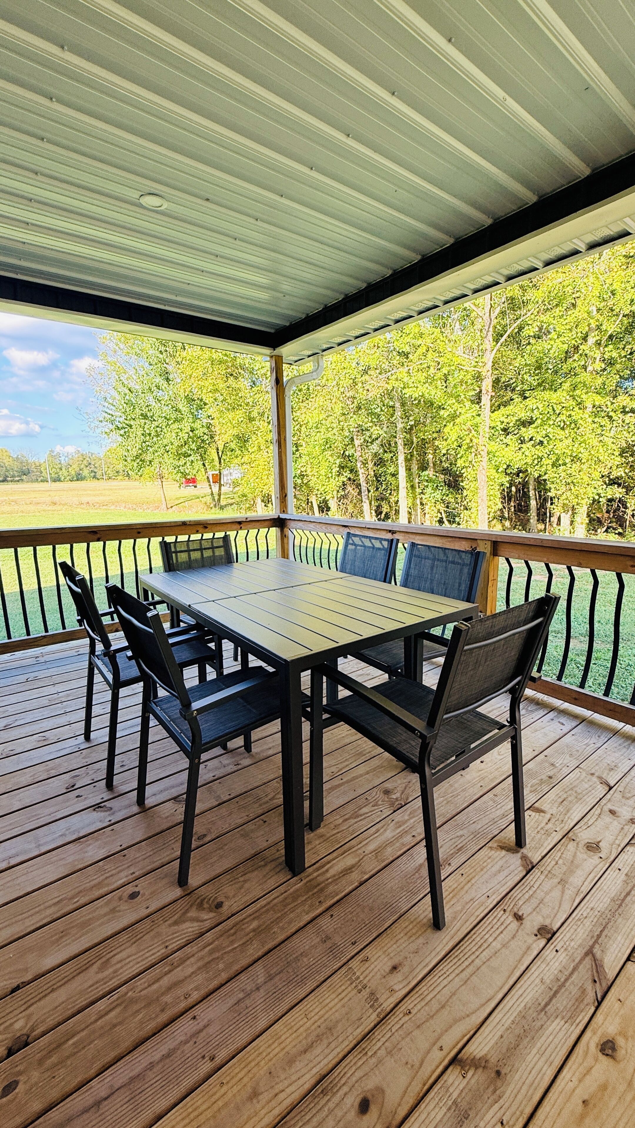 Een overdekt terras buiten met een houten tafel en stoelen, omgeven door een metalen hekwerk.