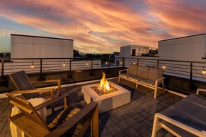 A rooftop terrace with a fire pit, seating, and a cityscape view at dusk.