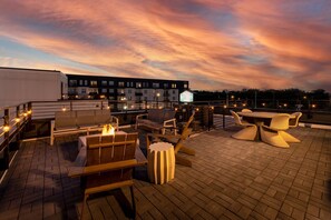 Terraza en la azotea con mobiliario exterior, una chimenea y vistas a edificios bajo un cielo al atardecer.