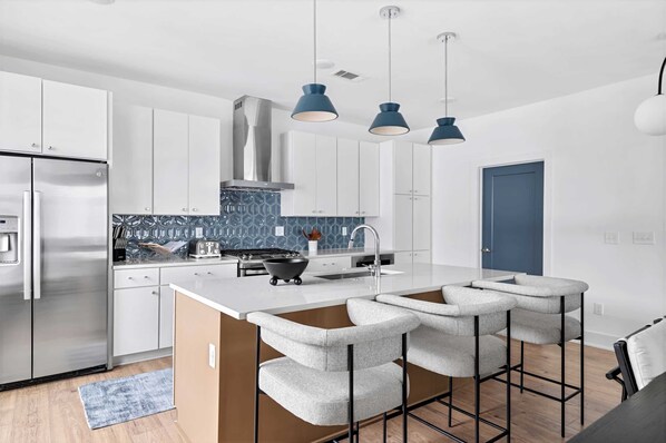 Et moderne køkken med hvide skabe, et rustfrit stål køleskab, blå backsplash og en central ø med barstole.