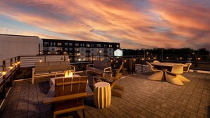 Terraço no telhado com área de estar ao ar livre, lareira e vista para edifícios sob um céu ao pôr do sol.