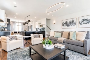 A modern living room with a sofa, two armchairs, a coffee table with plants, and a kitchen in the background.
