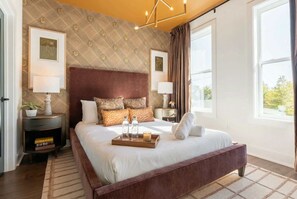 A spacious bedroom with a large bed, a nightstand with a lamp, a bedside table with a plant, and a window with curtains.