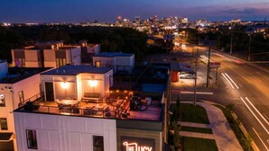 A modern building with a rooftop patio area, a sign reading "The Lucy," and a city skyline in the background.