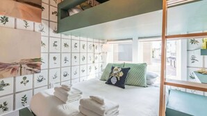 A neatly made bed with white linens and a decorative pillow, a stack of folded towels, and a view of the outdoors through a window.
