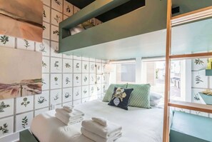 A neatly made bed with white linens and a decorative pillow, a stack of folded towels, and a view of the outdoors through a window.