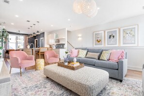 Una sala moderna con un sofá gris, sillones rosas, un otomán grande y un comedor con una cocina al fondo.