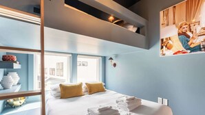 A modern hotel room with a large bed, two pillows, a bedside table with a lamp, and a wall-mounted shelf with decorative items.