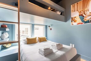 A modern hotel room with a large bed, two pillows, a bedside table with a lamp, and a view of the city through large windows.