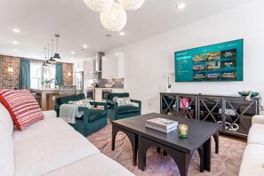 A modern living room with a sofa, armchairs, and a coffee table. A flat-screen TV is mounted on the wall displaying a menu.