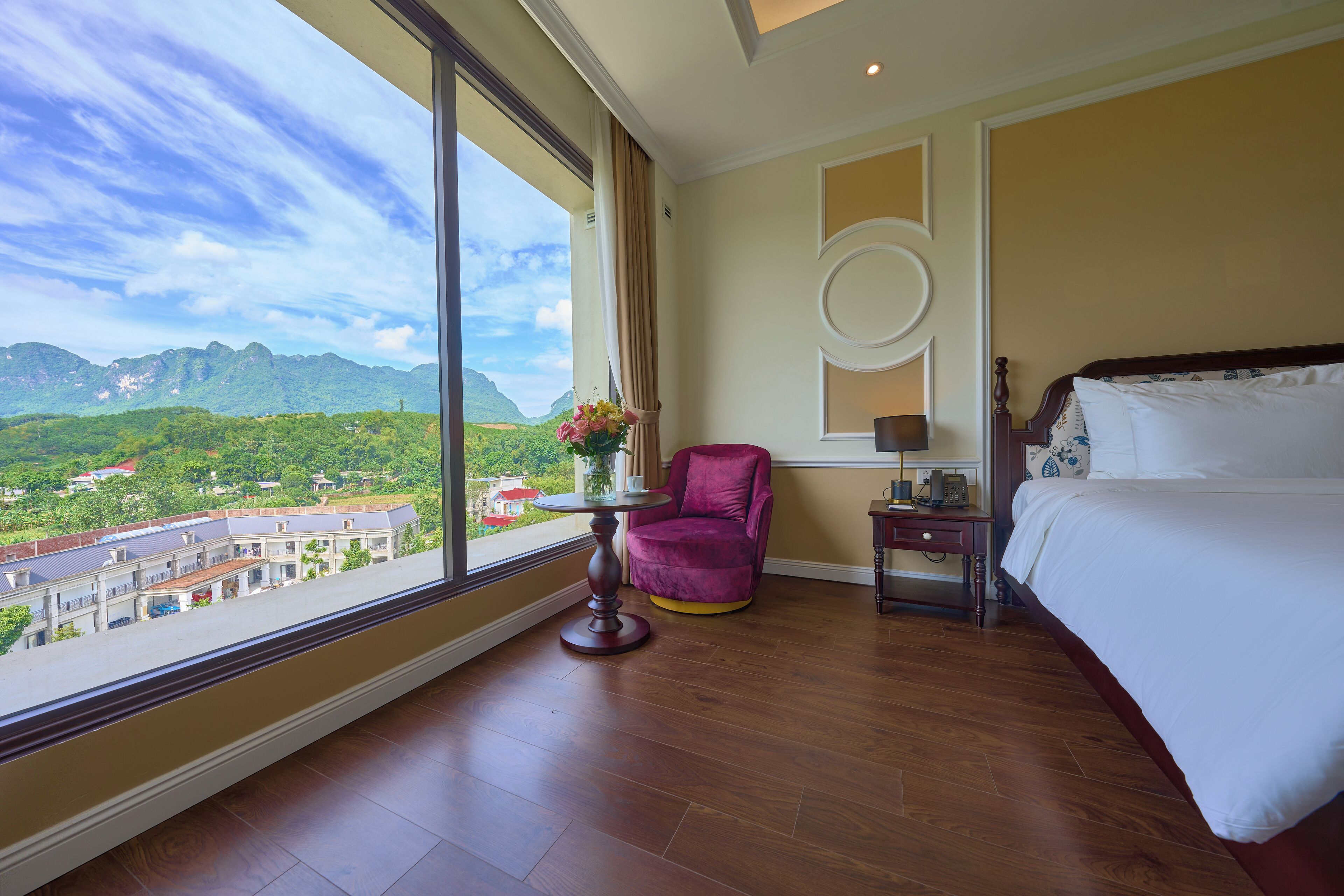 Une chambre d’hôtel avec une grande fenêtre, un lit, une chaise et une petite table sur laquelle est posé un vase de fleurs.