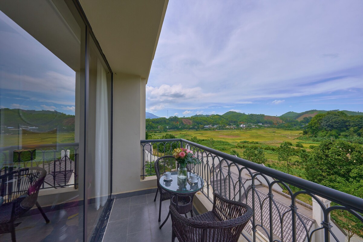 A balcony with wicker furniture and a table, overlooking a green landscape with hills.
