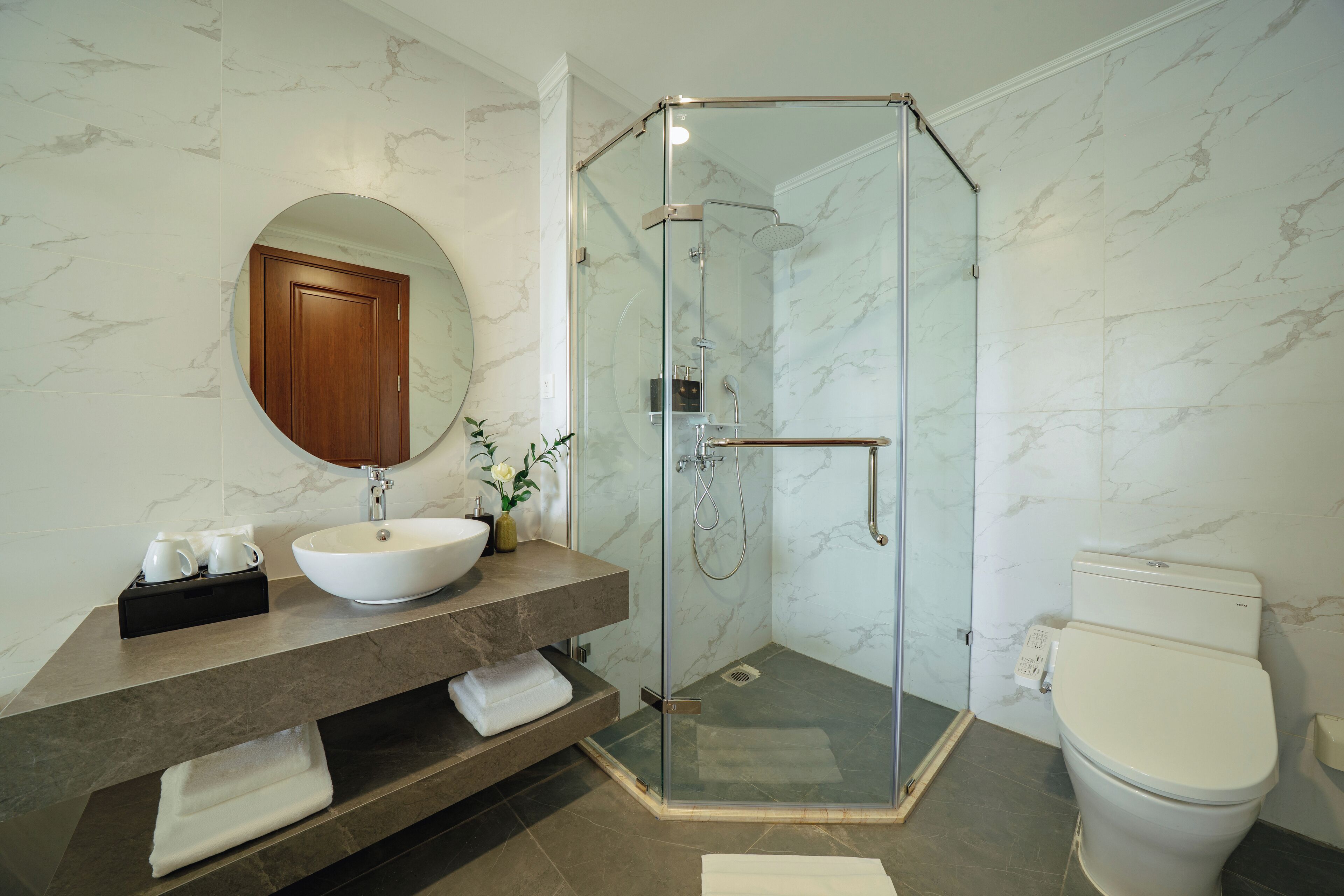 A modern bathroom with a glass-enclosed shower, a marble-tiled wall, a wooden vanity, and a white toilet.