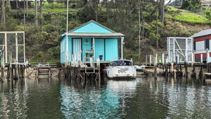 A turquoise wooden structure with a small deck, surrounded by calm water and docked boats. - Stylish Seaside Retreat with Spa (Narooma)