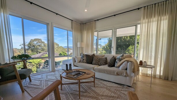 A spacious living room with a large white sofa, a wooden coffee table, and a view of a lush landscape through large windows. - Stylish Seaside Retreat with Spa (Narooma)