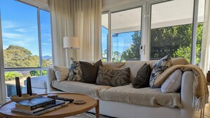 A modern living room with a beige sofa, a round wooden coffee table, and a view of greenery through large windows. - Stylish Seaside Retreat with Spa (Narooma)