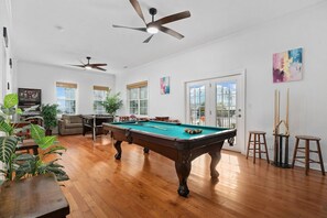 Une table de billard dans une pièce dotée d’un plancher en bois, de ventilateurs de plafond et d’une vue sur l’extérieur, visible à travers une porte vitrée.