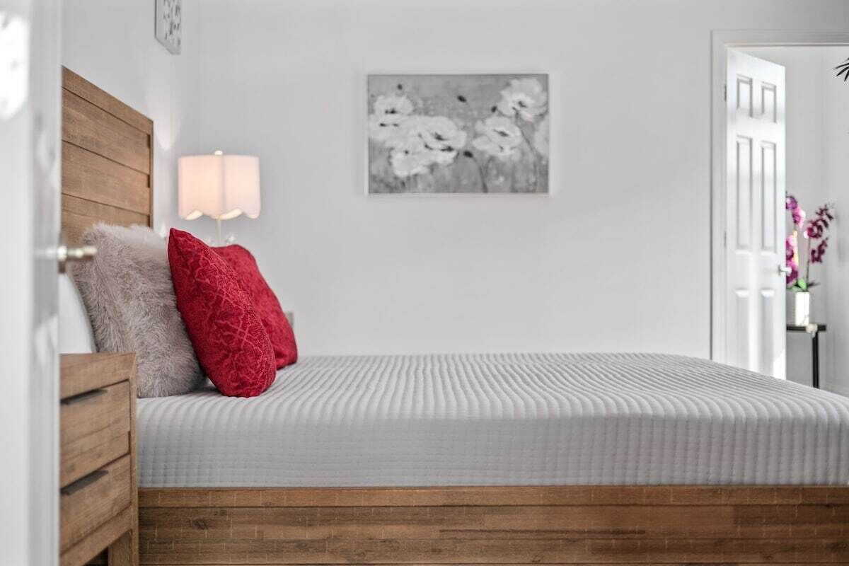 Ein Schlafzimmer mit einem Holzkopfende, einer weißen Bettdecke, einem roten Kopfkissen, einer Wandlampe und einem schwarz-weißen Blumenbild.