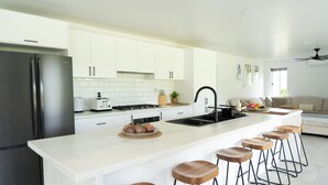 A modern kitchen with white cabinets, a stainless steel refrigerator, a central island with stools, and a dining area with a sofa and a ceiling fan. - Enua Tipani Pool Villa
• Quiet • Convenient location• Modern Finishes • Sleeps 8 (Rarotonga)