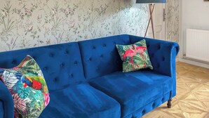 A blue sofa with floral and tropical patterned cushions, positioned in a room with floral wallpaper and a wooden parquet floor. - 1 bedroom accommodation in St Athan , near Cardiff (St Athan , near Cardiff)