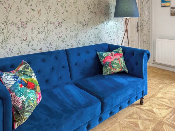 A blue sofa with floral and tropical patterned cushions, positioned in a room with floral wallpaper and a wooden parquet floor.