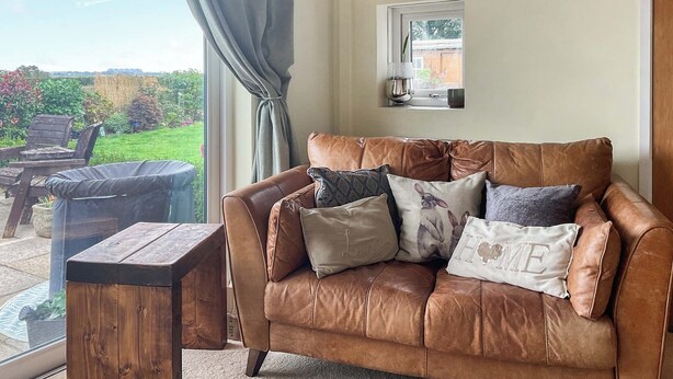 A brown leather sofa with multiple cushions, a wooden coffee table, and a view of a garden through a large window with a curtain. - 1 bedroom accommodation in St Athan , near Cardiff (St Athan , near Cardiff)