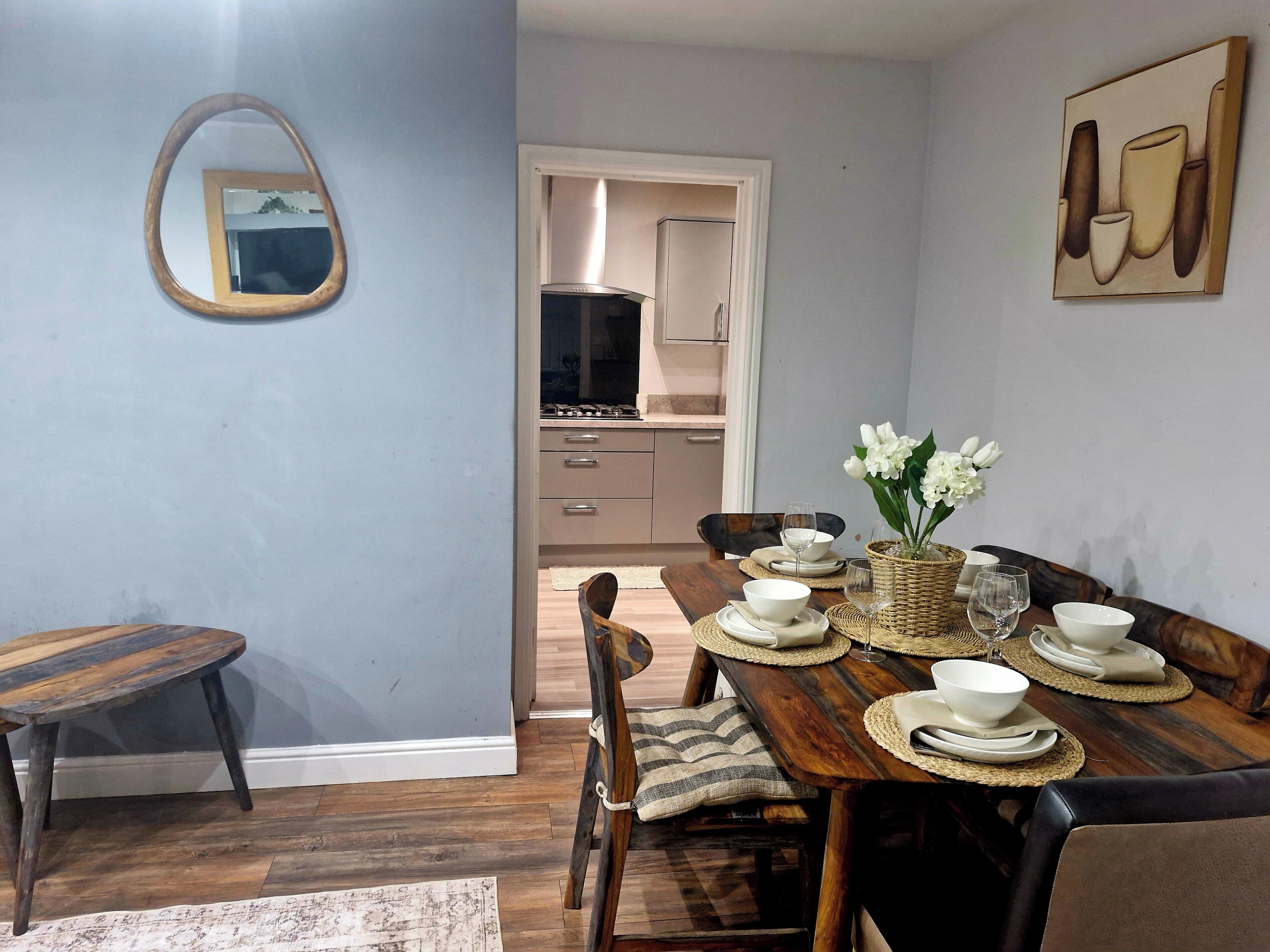 Un coin repas avec une table en bois dressée pour quatre personnes, un miroir et une cuisine en arrière-plan.