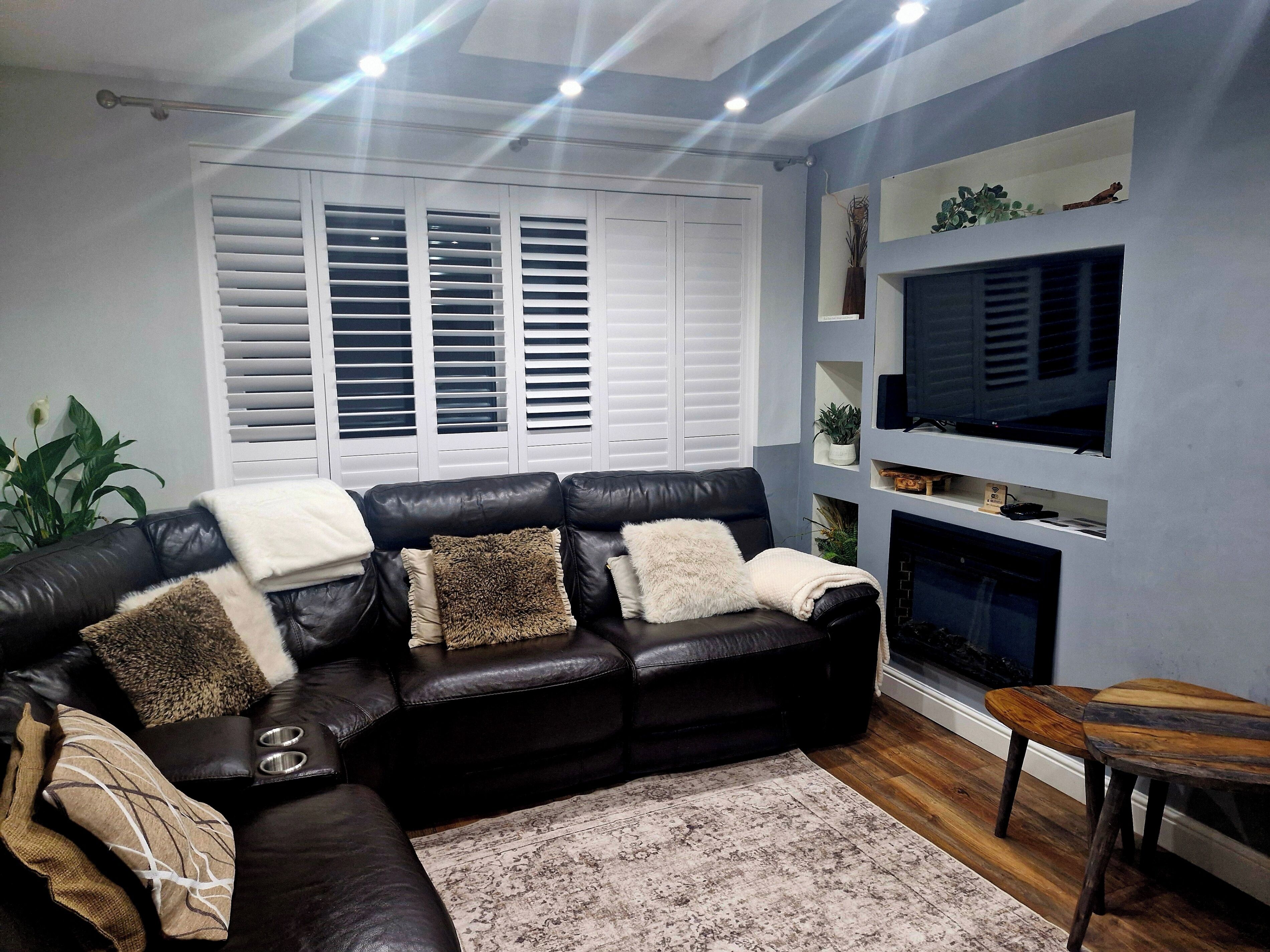 A living room with a black leather sofa, white shutters, a fireplace, and a TV mounted on the wall.