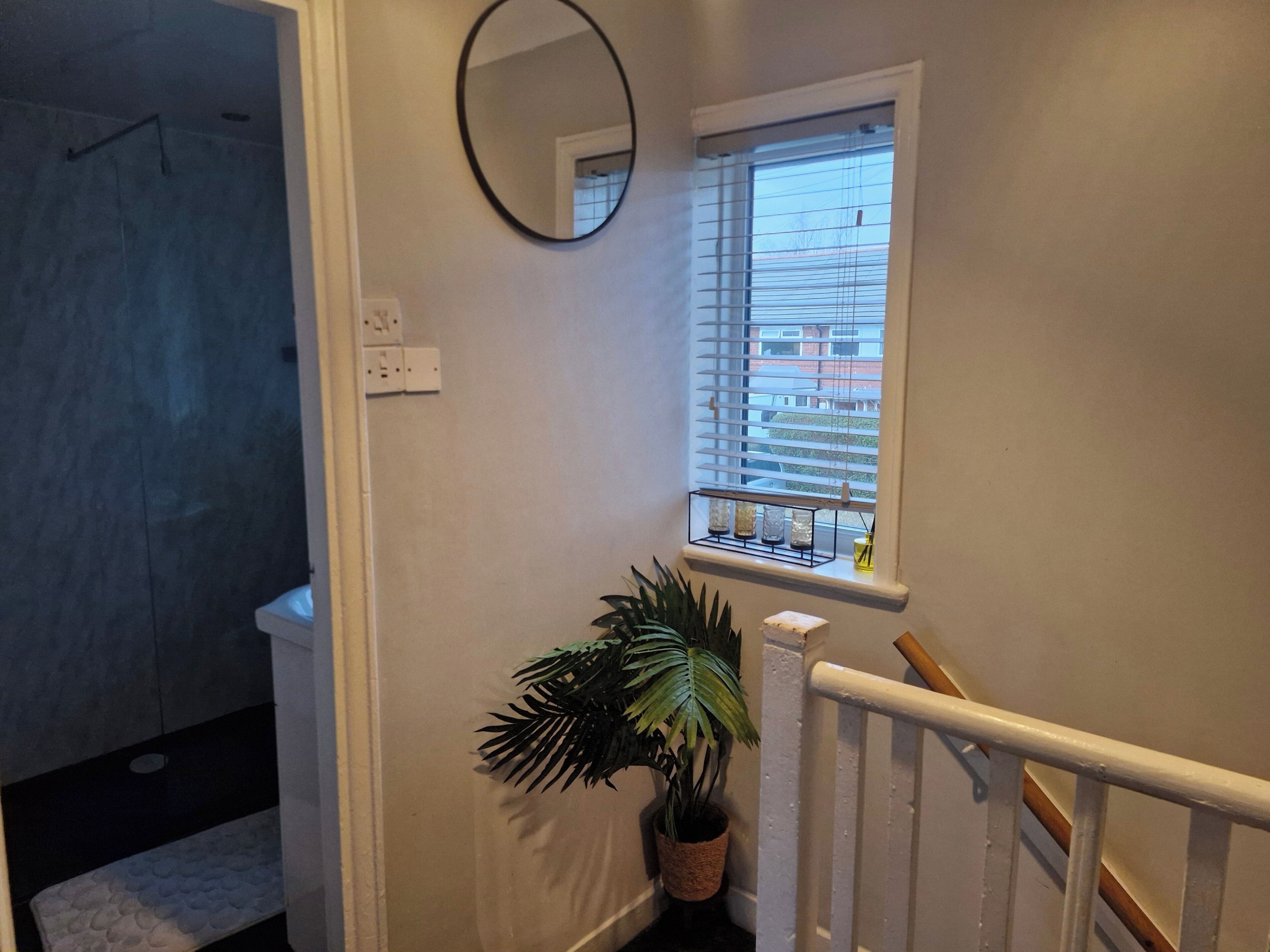 Un escalier avec une rampe blanche, une plante en pot, une petite fenêtre avec des stores et un miroir fixé au mur.