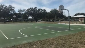 En utomhusbasketplan med en grön yta, en basketkorg och ett avskärmat sittområde.