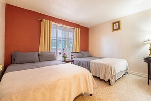Quarto de hotel com duas camas, janela com cortinas, uma mesinha com um vaso de flores e uma luminária em uma mesa de cabeceira.