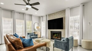 Een moderne woonkamer met een leren bank, een blauwe fauteuil en een poef. Een flatscreen televisie is boven een open haard bevestigd.