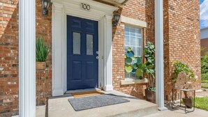 Een bakstenen huis met een donkerblauwe deur, het cijfer '100' boven de deur en een klein raam met decoratieve glaspanelen.