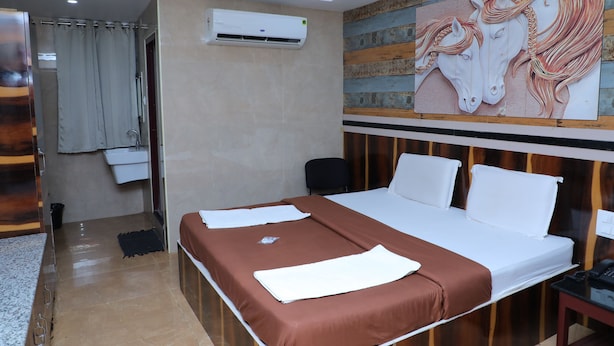 A hotel room with a bed, a desk with a phone, an air conditioning unit, a painting of horses, and a bathroom with a shower curtain. - SSR HOTEL (Srikalahasti)
