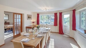 A dining room with a long wooden table set for six, a small side table with a lamp, and a window with red curtains. - 4 bedroom accommodation in Welshpool (Welshpool)