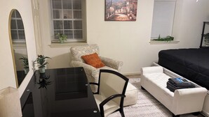 Een kleine leefruimte met een zwart bureau, een witte fauteuil met oranje kussen, een witte bank en een bed met zwart dekbed. Er zijn twee ramen met zonwering, een schilderij aan de muur en een plafondventilator.