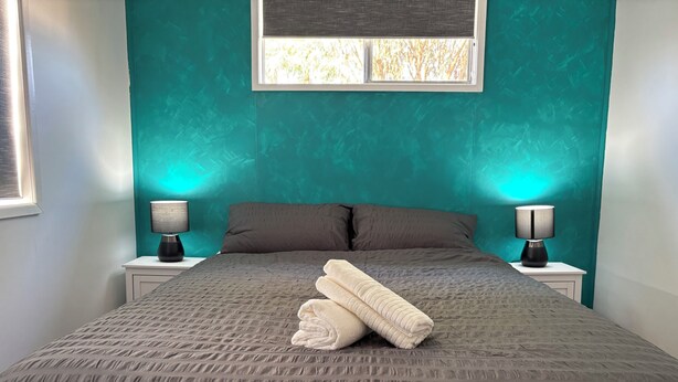 Une chambre avec des murs de couleur turquoise, une couvre-lit grise, deux oreillers, deux lampes de chevet et une fenêtre donnant sur des arbres.