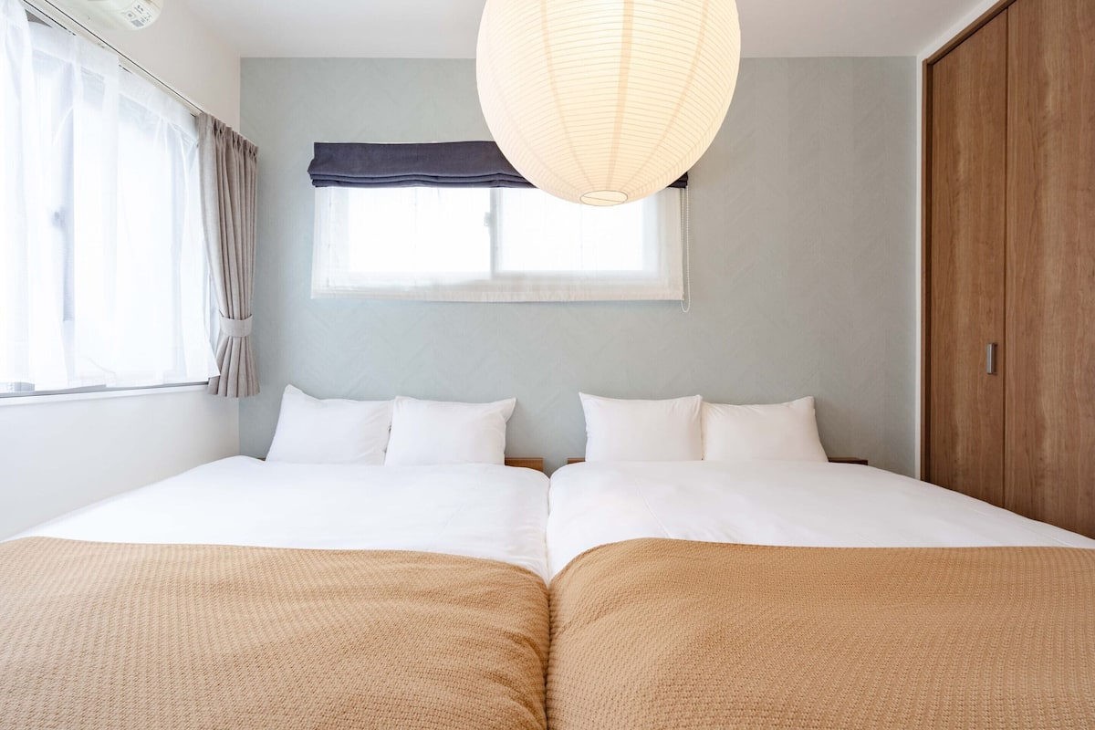 A double bed with white linens, a beige throw blanket, a window with curtains, and a hanging light fixture.