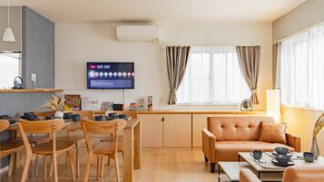A modern living room with a sofa, dining table, chairs, a flat-screen TV, and a window with curtains.