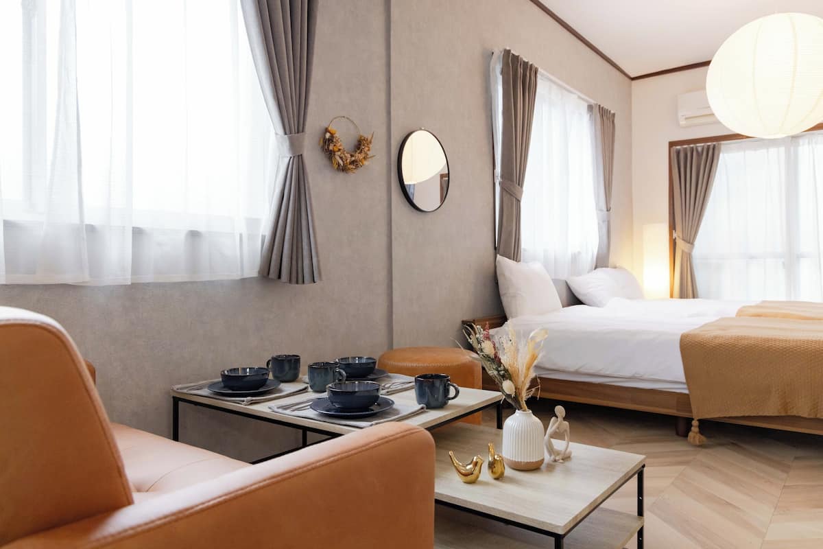 A modern hotel room with a bed, a dining table with chairs, a sofa, and a small table with decorative items.