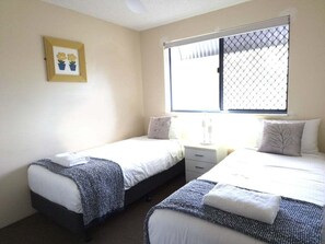A room with two beds, a window with a mesh screen, a bedside table with a lamp, and a framed picture on the wall.