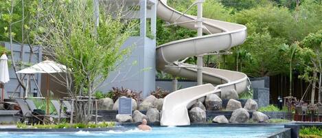A pool with a multi-level water slide, surrounded by rocks and greenery.