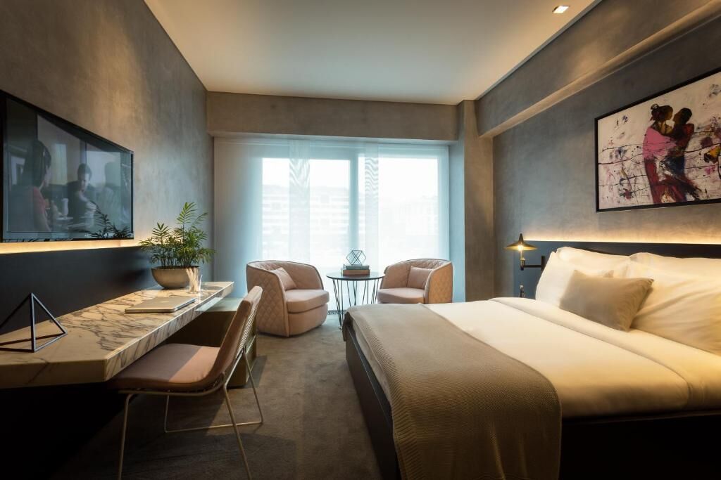 Une chambre d’hôtel moderne avec un grand lit, un bureau avec une chaise, une petite table avec deux chaises, une plante en pot et des œuvres d’art accrochées aux murs.